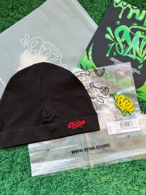 Svart mössa från SYNA med röd logga - Svart äkta mössa från SYNA med röd broderad graffiti-logga framtill. Tillverkad i mjuk bomull och polyesterblandning, enkel och clean design som passar streetwear-stilen. Mössan är stretchig och sitter skönt på huvudet.