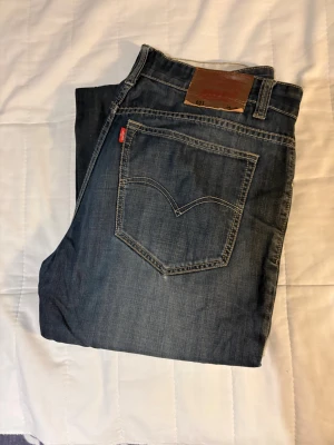 Levi's 501 mörkblå jeans straight fit - Säljer ett par klassiska Levi's 501 jeans i mörkblå tvätt med snygga slitningar. Modellen har rak passform, fem fickor och tydliga kontrastsömmar. Jeansen har normal midja och är tillverkade i robust denim med Levi's logga på bakfickan. W 34 L 32