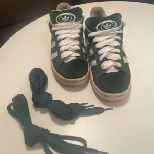 Adidas Campus 00s sneakers mörkgröna - Adidas Campus sneakers i mörkgrön mocka med vita klassiska stripes och logga på tungan och hälen. Platt gummisula i brun färg och rund tå. Skorna har vita snören och extra gröna snören medföljer. Snygg retrostil och låg profil. Dessa är i storlek 41 1/3 men själv har jag storlek 42 och dem passar som bäst nu. Använda men i fortfarande mycket bra skick, Mockan i fortsatt bra skick. Ett litet hål i höger häl kappa inget som känns dock.  Ny pris 1499kr