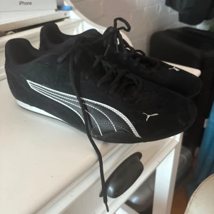 Svarta Puma sneakers med vit logga - Säljer ett par svarta Puma sneakers med klassisk vit logga och sidospår. Skorna har snörning och är tillverkade i mocka med platt sula och rund tå. Endast använda 2 gånger på gymmet, men passar att ha utomhus också .