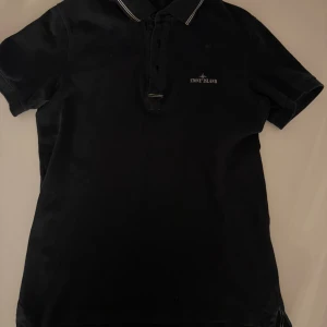 Svart piké från Stone Island - Svart pikétröja från Stone Island med klassisk krage och vita ränder på kragen. Tröjan har knappslå framtill och broderad logga på bröstet. Perfekt för en clean och stilren look. Använd fåtal gånger, nypris 1800 pris kan diskuteras vid snabb affär!! 