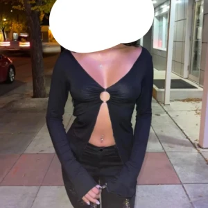 Svart croppad omlottopp med ring - Säljer en svart långärmad topp med djup v-ringning och en metallring framtill som håller ihop tyget. Toppen är croppad och har vågiga kanter nertill. Ganska stretchig så passar M oxå