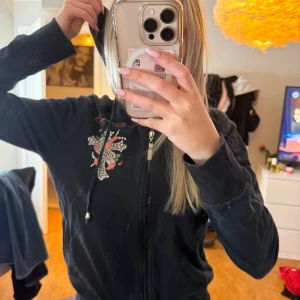 Svart Ed Hardy zip up - Svart hoodie från Ed Hardy med stort färgglatt tryck av kors, hjärta och blommor på ryggen samt mindre tryck fram. Dragkedja framtill, huva med snörning och långa ärmar. Materialet känns mjukt och skönt, perfekt för chill dagar.