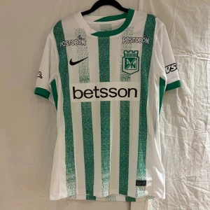 Atlético Nacional fotbollströja Morelos #9 - Säljer en  Atlético Nacional fotbollströja med Morelos och nummer 9 på ryggen. Tröjan är vit med gröna vertikala ränder, har korta ärmar och gröna muddar. Märkt med klubbens emblem, sponsorloggor och texten 'El Verde de la Montaña' i nacken. Tillverkad i lätt polyester.