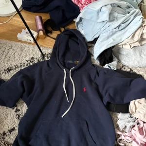 Marinblå hoodie från Polo Ralph Lauren - Säljer en marinblå hoodie från Polo Ralph Lauren med vit snörning och klassisk röd logga på bröstet. Tröjan har huva, känguruficka och är långärmad. Perfekt för chill dagar och enkel att matcha med jeans eller joggers.