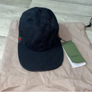 Svart keps från Gucci med GG-mönster - Svart keps från Gucci med klassiskt GG-monogram över hela kepsen och ikoniska gröna och röda ränder på sidan. Justerbar rem baktill i svart skinn. Kepsen är tillverkad i bomull och har märkets logga på insidan samt etikett och tag kvar.✅