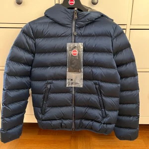 Mörkblå Colmar pufferjacka med huva - Snygg mörkblå pufferjacka från Colmar med huva och två sidofickor med dragkedja. Jackan har quiltad design och hel dragkedja framtill. Perfekt för kalla dagar och har en clean look som funkar till många stilar. Nypris är 5700kr på kidsbrandstore