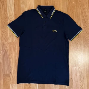 Marinblå pikétröja från BOSS - Snygg marinblå pikétröja från BOSS med gul logga på bröstet. Kragen och ärmsluten har vita och gula ränder som ger en sportig touch. Klassisk passform och korta ärmar, perfekt till jeans eller shorts.