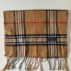 Rutig halsduk från Burberry - Snygg klassisk halsduk från Burberrys i 100% lammull. Beige botten med rutigt mönster i svart, vitt och röda linjer. Mjukt material och fransar i ändarna. 
