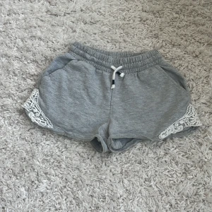 Grå shorts med spets från Lindex - Snygga grå shorts från Lindex med vit spetsdetalj på sidorna och bekväm snörning i midjan. Mjukt bomullsmaterial och elastisk midja gör dem perfekta för chill dagar. Passar dig som gillar enkel stil med en twist.