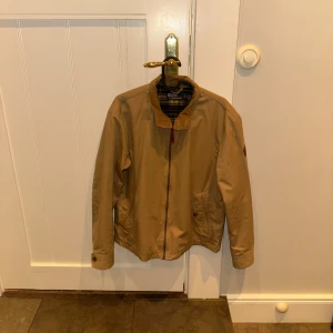 Beige jacka från Polo Ralph Lauren - Snygg beige jacka från Polo Ralph Lauren med klassisk krage i manchester och rutigt foder i blått och gult. Jackan har dragkedja framtill, knappar vid ärmslut och fickor med knappstängning. Materialet är 100% bomull Ventile, vädertålig och perfekt för höst och vår.