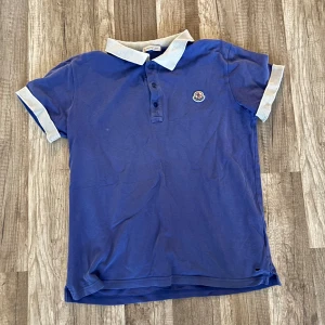 Blå Moncler pikétröja med vit krage - Snygg blå pikétröja från Moncler med vit kontrastkrage och ärmslut. Klassisk Moncler-logga på bröstet och broderad Moncler-text på baksidan av kragen. Tröjan har knappar framtill och är tillverkad i mjuk bomull.