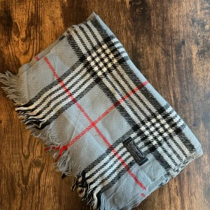 Burberry halsduk - Klassisk Burberrys-halsduk i 100% lammull, tillverkad i England. Mjukt och varmt material i ikoniskt rutmönster. Passar både dam och herr. I mycket bra skick små fläckar eller utan hål – perfekt för höst och vinter. 
