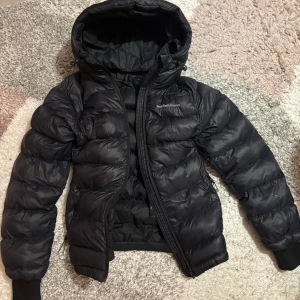 Svart Peak Performance dunjacka - Svart dunjacka från Peak Performance med huva och dragkedja framtill. Jackan har puffad design, ribbade muddar vid ärmslut och två sidofickor med dragkedja. Perfekt för kalla dagar och har en stilren look som funkar till det mesta. Använd en vinter , som ny köpt för 3499:-