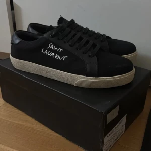 Svarta Saint Laurent sneakers - Svarta sneakers från Saint Laurent med vit handskriven logga på sidan. Skorna har snörning, rund tå och platt vit sula. Ovandelen är i mocka och skinn, vilket ger en lyxig och stilren vibe. Perfekta för dig som gillar exklusiva streetwear och snygga detaljer.