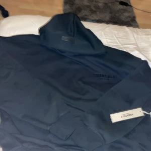 Svart Essentials Fear of God hoodie - Svart hoodie från Essentials Fear of God med stor logga på bröstet och patch på huvan. Tröjan har en klassisk oversized passform, mjukt material och huva. Perfekt för dig som gillar streetwear och vill ha en clean look.