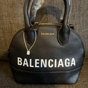 Zara väska -  svart handväska med vit logotyp framtill och guldfärgade detaljer. Väskan är tillverkad i strukturerat skinn och har rundad form med två handtag och dragkedja upptill. 