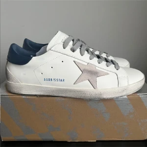 Golden Goose vita sneakers med blå detalj - Golden Goose sneakers i vitt skinn med grå mockastjärna på sidan, grå snörning och mörkblå hälkappa. Snygg vintage-look med slitna detaljer och platt sula. Perfekta för dig som gillar en avslappnad men trendig stil.