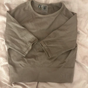 Beige ribbad tröja från Stronger - Snygg beige ribbad tröja från Stronger med trekvartsärm och rund hals. Tröjan har ett diskret broderat Stronger-logo på nederkanten och är gjord i mjuk polyester som känns skön mot huden. Perfekt för chill eller träning.