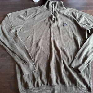 Grön half zip tröja US Polo Assn - Grön långärmad tröja från US Polo Assn med half zip och ståkrage. Tröjan har ribbade muddar vid ärmslut och nederkant samt en broderad logga på bröstet. Perfekt för dig som gillar klassisk stil med sportig touch.