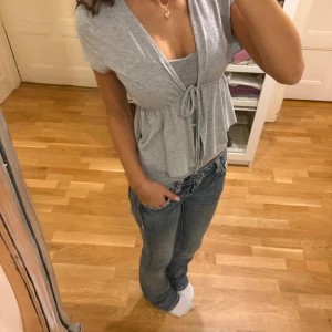 Grå omlottopp med knyt från Gina Tricot - Supersöt grå omlottopp från Gina Tricot i mjuk bomull. Toppen har korta ärmar, v-ringning och ett knyt i midjan som ger en snygg siluett. Perfekt att styla med jeans eller kjol för en chill look. Passar dig som gillar enkel och clean stil.