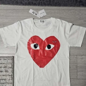 CDG t shirt - Säljer en vit t-shirt från CDG PLAY med stort rött hjärta med ögon tryckt på bröstet och ett mindre broderat hjärta ovanpå. Klassisk rund hals och korta ärmar. Materialet är mjuk bomull, perfekt för en clean och cool look.