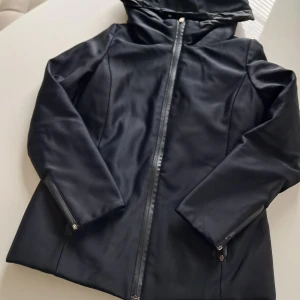 Svart pufferjacka från Weekend - Svart pufferjacka från Weekend med huva och snygga dragkedjor både framtill och vid ärmsluten. Jackan har en stilren design och är tillverkad i syntetmaterial med quiltad insida för extra värme. Perfekt för kalla dagar och har en normal passform.