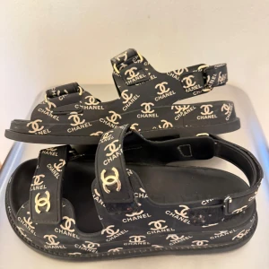 Äkta! Svarta Chanel sandaler med logga  - Äkta! Slutsålda coola klassiker!!Svarta sandaler från Chanel med platt sula och rund tå. Sandalerna har flera remmar med justerbart spänne och är täckta av vita Chanel-loggor i ett mönster. Materialet är syntet och detaljerna är i guld. Perfekt för dig som gillar statement pieces och lyxig streetstyle.