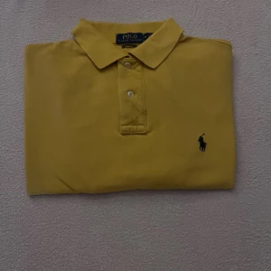 Gul pikétröja från Polo Ralph Lauren - Klassisk gul pikétröja från Polo Ralph Lauren i bomull med broderad marinblå logga på bröstet. Tröjan har krage, knäppning med två knappar och en clean, tidlös look. Perfekt för dig som gillar stilrena och sportiga vibes.
