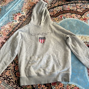 Grå hoodie från GANT med logga - Säljer en grå hoodie från GANT med broderad logga och sköld på bröstet. Tröjan har huva med snörning och en stor magficka. Perfekt för dig som gillar klassisk och sportig stil.
