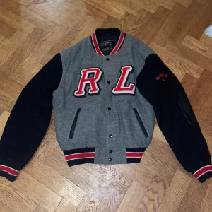 Grå och svart bomberjacka Polo Jeans  - Snygg bomberjacka från Polo Jeans Co. med grå kropp och svarta ärmar, röda broderade RL-bokstäver och detaljer. Jackan har ribbade muddar med röd och vit rand, knappar framtill och svarta skinnlappar på armbågarna. Materialmix av ull och viskos. Det är en väldigt ovanlig jacka som inte görs längre. 