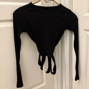 Svart ribbad omlottopp med knyt i ryggen - Säljer en svart ribbad omlottopp med långa ärmar och snygg knytning baktill. Toppen har en croppad passform och är tillverkad i mjukt, stretchigt material som sitter skönt mot kroppen. Använd endast en gång!🥰