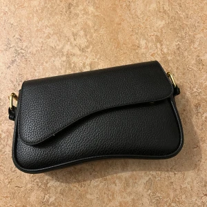 Svart axelväska i skinn med gulddetaljer - Stilren svart axelväska i skinn med snygg struktur och guldfärgade metalldetaljer. Väskan har ett lock med magnetknäppning och ett innerfack med dragkedja. Perfekt storlek för det viktigaste och en modern, kurvad form.