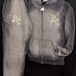 Decrease Tracksuit – 100% bomull, storlek S - Upplev stil och komfort med detta exklusiva set från Decrease – perfekt för vardag eller streetwear! Setet består av mörkgrå hoodie och matchande byxor med vit broderad logga. 100 % bomull, mjukt och slitstarkt. Hoodie med drake på huvan och broderier fram/bak. Byxor med resår, dragsko och flera fickor. Nytt och oanvänt – hög kvalitet! Kan skickas eller mötas i Stockholm. 