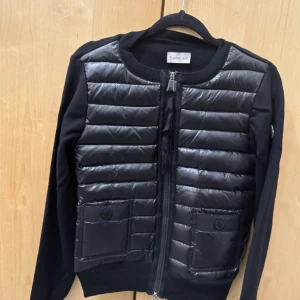 Svart quiltad cardigan jacka Moncler - Svart cardigan jacka från Moncler med quiltad front i blankt material och stickade ärmar och rygg. Jackan har två fickor med knapp framtill, rund hals och dragkedja. Moncler-logga på ärmen ger extra premiumkänsla. Perfekt för dig som gillar stilren design.