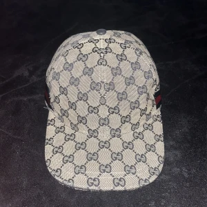 Gucci keps  - Säljer en snygg keps från Gucci med klassiskt GG-monogram i blått på grå botten. Kepsen har böjd skärm och ikoniska röda och blå ränder på sidan. Tillverkad i bomullscanvas med mörkblått innerfoder och märkets logga invändigt.