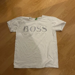Vit BOSS t-shirt med tryck - Vit t-shirt från BOSS med stort svartmönstrat BOSS-tryck på bröstet. Klassisk passform med rund hals och korta ärmar. Tillverkad i mjuk bomull, perfekt för en clean och stilren look.