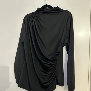 Svart långärmad polotopp från SHEIN - Svart polotopp från SHEIN Curve med lång ärm och cool drapering på ena sidan. Toppen har en hög krage och är gjord i mjukt syntetmaterial som känns skönt mot huden. Perfekt för dig som gillar stilrena plagg med en twist. Passar för storlek L-XL också