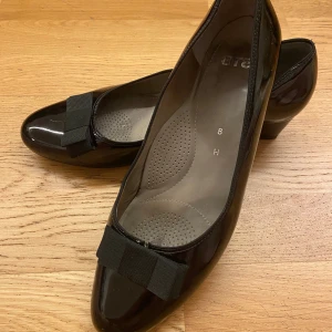 Svarta lackpumps med rosett - Klassiska svarta pumps(storlek 8=ish 41) i lack med rund tå och låg klack. Har en bekväm innersula och ÄR vidklippt (bild 5) för att fötter med större skolyft ska vara bekväma! Perfekta för dig som gillar en tidlös och elegant stil. 