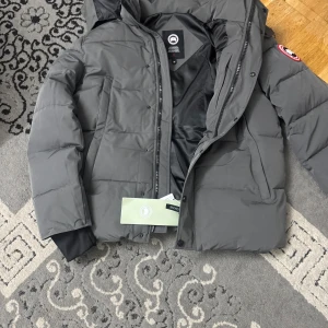 Mörkgrå pufferjacka från Canada Goose - Säljer en mörkgrå pufferjacka från Canada Goose med huva och klassisk logga på ärmen. Jackan har dragkedja och knappar framtill, ribbade muddar och två sidofickor. Fylld med dun och fjäder för extra värme, perfekt för kalla dagar. Materialet är polyester och dun. Priset kan diskuteras. Vid snabba affärer.