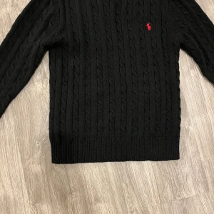 Svart kabelstickad tröja Polo Ralph Lauren - Svart kabelstickad tröja från Polo Ralph Lauren med klassisk röd broderad logga på bröstet. Tröjan har rund halsringning, långa ärmar och ribbade muddar. Perfekt för dig som gillar stilren och tidlös design.