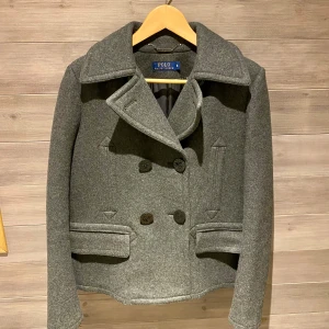 Grå skepparkavaj från Polo Ralph Lauren - Snygg grå skepparkavaj (kort kappa) från Polo Ralph Lauren med klassisk dubbelknäppning och bred krage. Jackan har två framfickor med lock och är tillverkad i mjuk men slitstark ull som ger en stilren och tidlös look. Innerfodret är silkeslikt och svalt. Perfekt för dig som gillar en clean och sofistikerad stil.