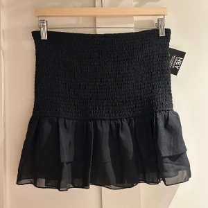 Svart smockad volangkjol från Nelly - Supersnygg svart kortkjol från Nelly med smockad midja och dubbla volanger nedtill. Kjolen har en luftig och lätt känsla, perfekt för att skapa en cool och trendig look. Materialet är mjukt och följsamt, och volangerna ger extra rörelse och stil.