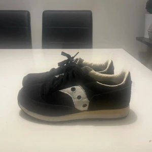 Svarta Saucony sneakers i mocka och textil - Säljer ett par svarta Saucony sneakers i storlek 43. Skorna har klassisk retrostil med vit kontrastdetalj på sidan, snörning och platt sula. Ovandelen är i mocka och textil, med gummisula för bra grepp. Perfekta för dig som gillar stilrena och sportiga sneakers. Och de passar perfekt för vintern också