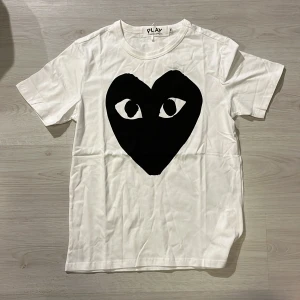 Vit t-shirt Comme des Garçons Play - Säljer en vit t-shirt från Comme des Garçons Play med det ikoniska svarta hjärtat med ögon tryckt på bröstet. T-shirten har rund hals och korta ärmar, tillverkad i mjuk bomull. Perfekt statement-plagg för dig som gillar streetstyle.