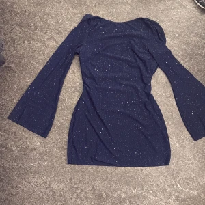 Glittrig marinblå kortklänning - Snygg marinblå kortklänning med glittrigt mönster och långa ärmar. Klänningen har en öppen rygg med snörning som ger en cool detalj. Materialet är mjukt och stretchigt, perfekt för att sticka ut. Passformen är slim och klänningen har en elegant siluett.