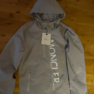 Grå vindjacka från Moncler - Snygg grå vindjacka från Moncler med huva och stor logotyp längs framsidan. Jackan har dragkedja, snörning vid halsen och en klassisk Moncler-patch på ärmen. Perfekt för dig som vill ha en clean och stilren look.