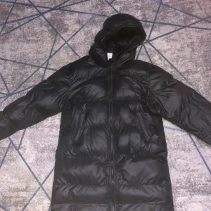 Svart lång pufferjacka från Nike XL - Svart pufferjacka från Nike i storlek XL med Therma-Fit teknologi. Jackan är lång, har huva och dragkedja framtill. Materialet är syntet och jackan har ett stilrent Nike-logga på bröstet. Perfekt för kalla dagar när du vill hålla värmen och se chill ut. Det finns ett litet hål på baksidan men man märker inte det så mycket.