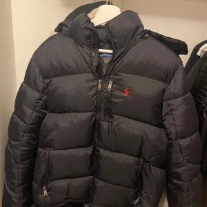 Svart pufferjacka från Ralph Lauren - Svart pufferjacka från Ralph Lauren med röd broderad logga på bröstet. Jackan har huva, flera dragkedjor framtill och är vadderad för extra värme. Perfekt för kalla dagar och har en klassisk, sportig look. 
