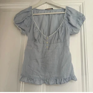 Ljusblå blus med puffärm och volang - Supersöt ljusblå blus i linne med puffärmar, djup v-ringning och tre små knappar framtill. Blusen har volangkant nertill och dekorativ spets längs urringningen. Perfekt för en somrig look!